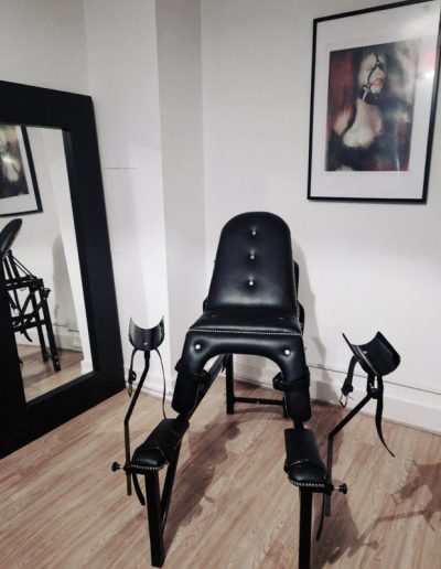 Elegant black BDSM chair with stirrups in a dimly lit room, framed art on the wall, mirror reflection visible.