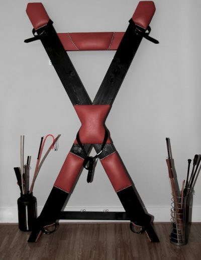 Black and red St. Andrew's cross in a room with various tools and gear on either side.
