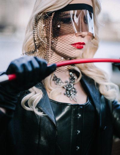 Glamorous woman in stylish black attire, visor, and veil, holding a red stick, exuding a mysterious vibe.
