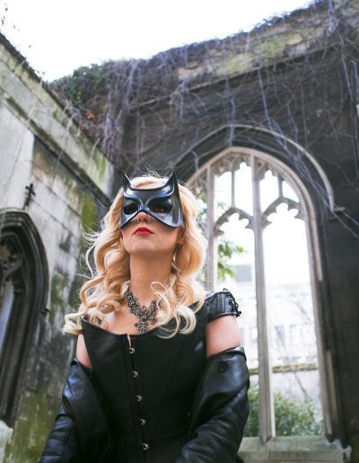 Woman in a black cat mask and costume stands in a gothic stone ruin with ivy-covered walls.