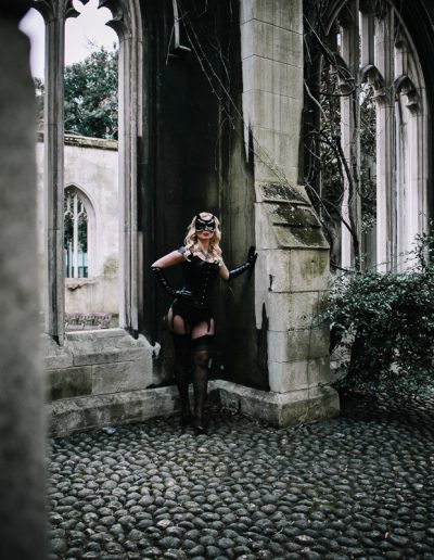 Woman in stylish outfit leans against Gothic archway in mysterious, ivy-covered stone ruin setting.