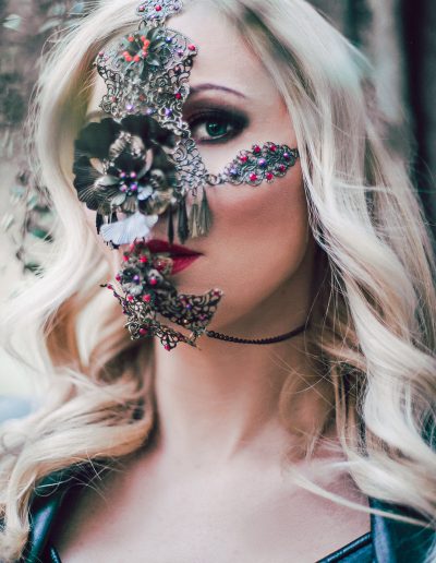 Woman wearing an intricate metal face mask with floral design and gemstones, set in a forest background.