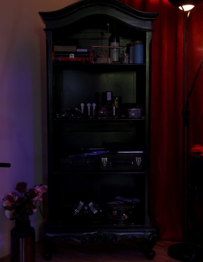 Dimly lit shelf with various decorative items, books, and wine against a red curtain backdrop.