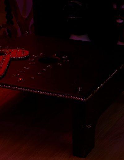 Black and red bondage table with metallic studs and wooden floor background, dimly lit room.