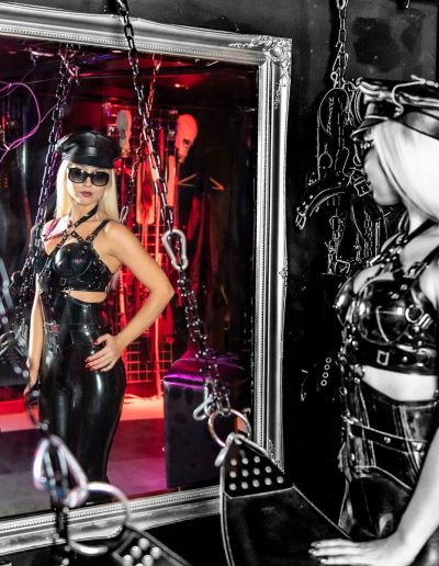 Woman in leather outfit and cap looking at herself in a mirror, surrounded by chains in a dimly lit room.