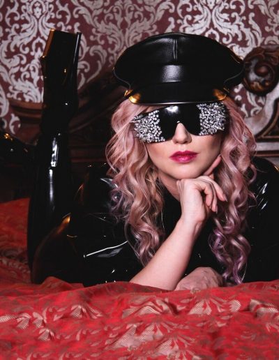 Woman with studded sunglasses and black outfit lying on a bed with red patterned sheets.