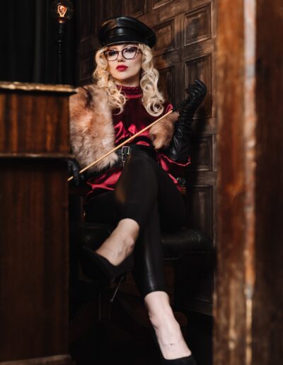 Blonde woman in vintage outfit with fur and leather, sitting confidently in stylish room with warm lighting.