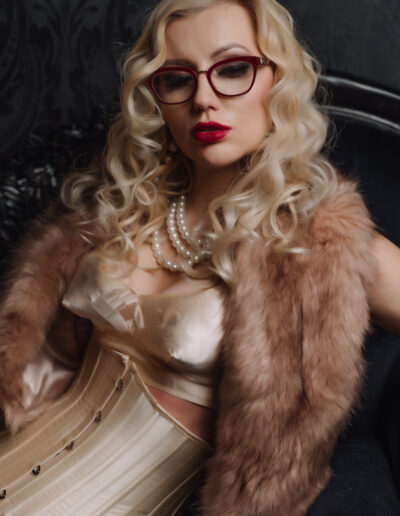 Stylish woman in glasses with curly blonde hair, wearing pearls, corset, and fur shawl in elegant setting.