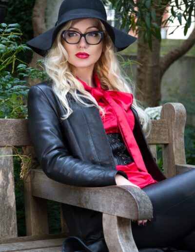 Fashionable woman in a black hat and leather jacket sitting on a bench, surrounded by greenery.
