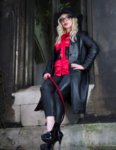Fashionable woman in black coat and hat with red blouse, sitting on steps in a gothic setting.